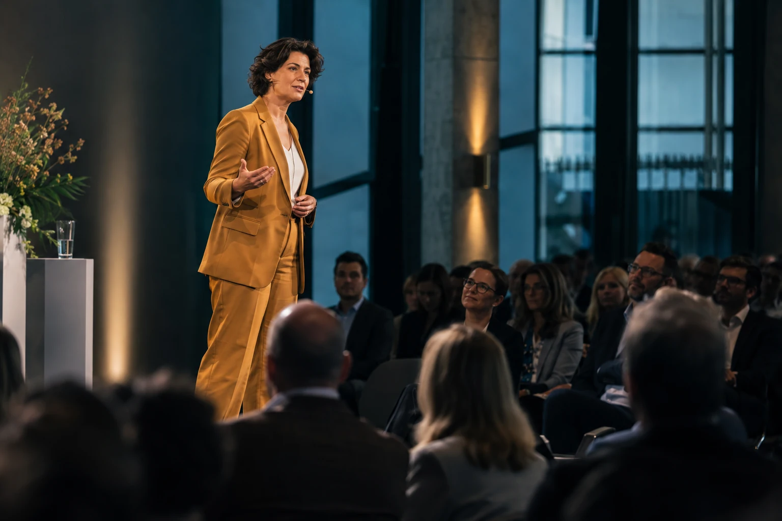 Eine Speakerin im gelben Anzug spricht vor dem Publikum einer Konferenz.